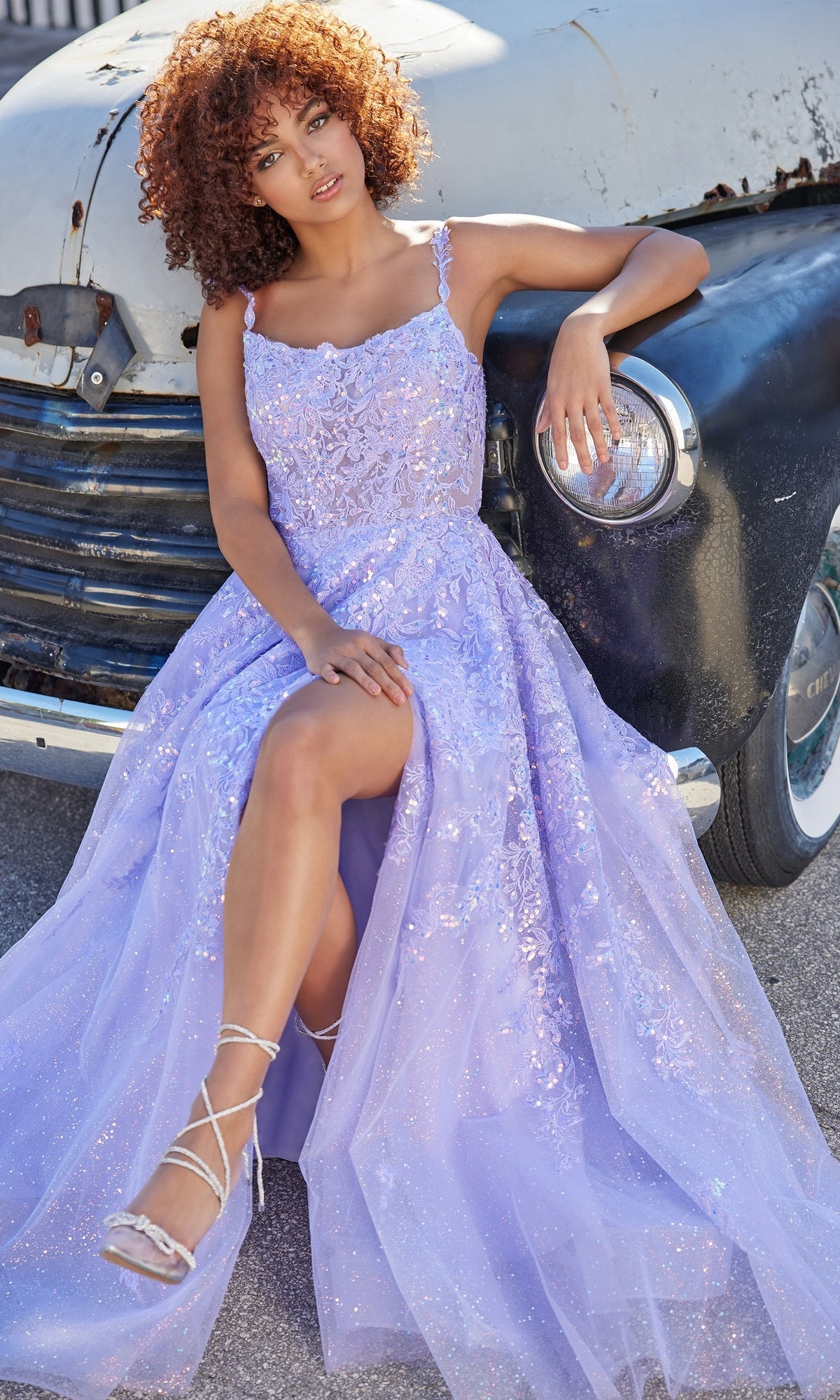 A woman wearing the Glitter-Tulle Ellie Wilde Long Prom Dress EW35114 sits on a vintage car bumper, showing one leg and strappy heels, while looking toward the camera.